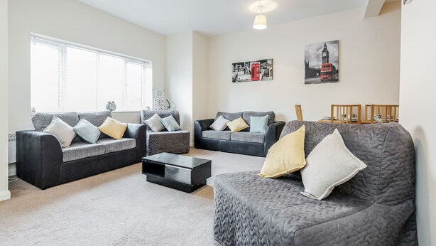 A living room with two grey sofas, a coffee table, and a TV. There are two windows with blinds, a painting of a red double-decker bus, and a clock on the wall.