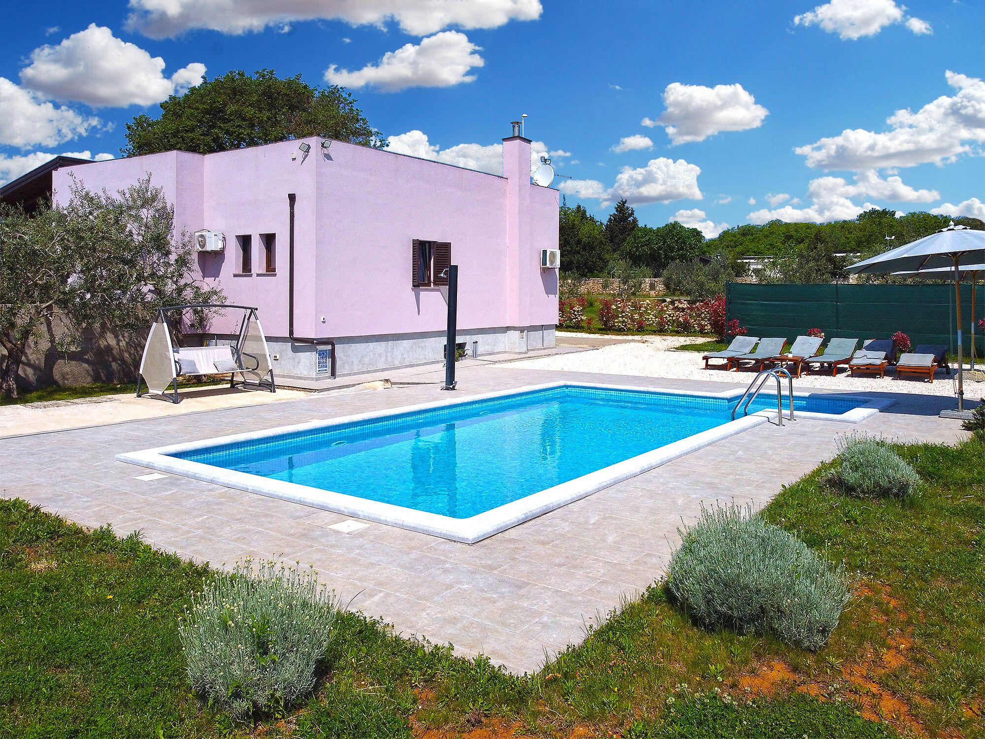 A swimming pool with a pink building in the background, surrounded by greenery and a clear sky.