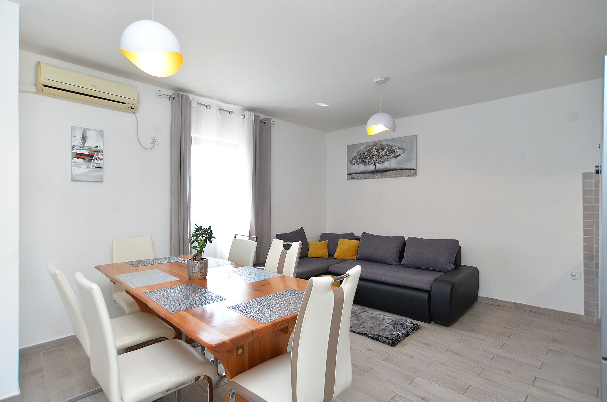 Una sala de estar moderna con una mesa de comedor de madera, sillas blancas, un sofá gris con cojines amarillos y una alfombra gris. Se observan dos lámparas colgantes, un aire acondicionado de pared y un cuadro de un árbol.