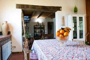A dining area with a table covered in a patterned tablecloth, a bowl of fruit, and a rustic wooden ceiling.
