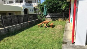 A grassy outdoor area with a picnic table and benches, a green umbrella, and a red pipe near a building. - White House (Onna)