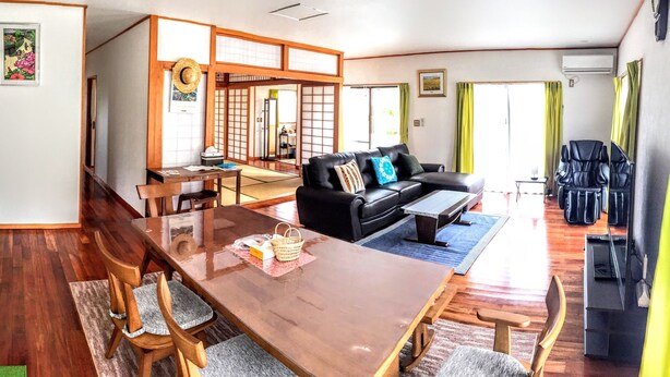 A traditional Japanese-style living room with tatami flooring, a black leather sofa, a wooden dining table with chairs, and a sliding shoji screen. - White House (Onna)