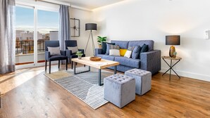 Una sala moderna con un sofá gris, una mesa de centro y un taburete gris. Hay una lámpara de pie y una mesita con una planta.