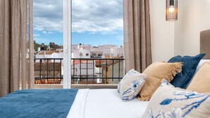 Un dormitorio con una cama grande, vista a edificios a través de una ventana y un balcón con barandilla.