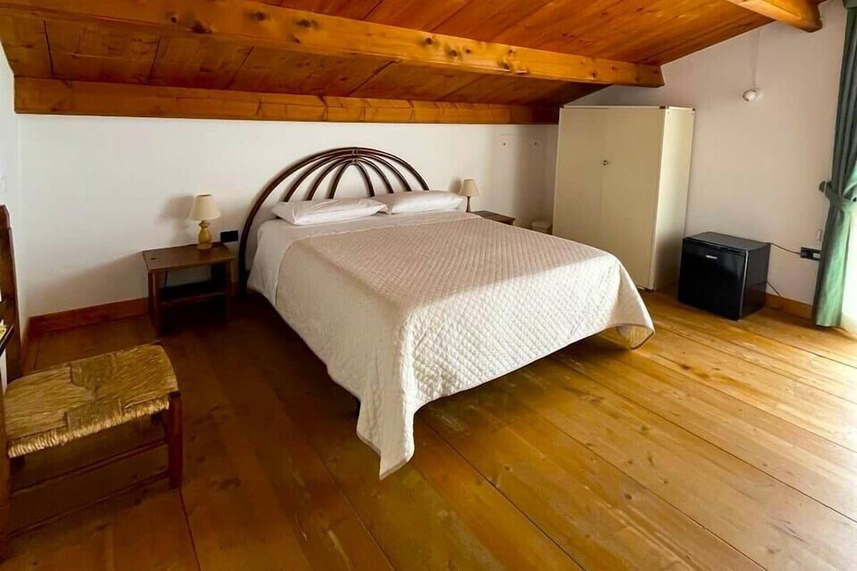 Une chambre confortable avec un plafond en bois, un lit recouvert d’une couette blanche, une table de chevet avec une lampe et un petit banc.