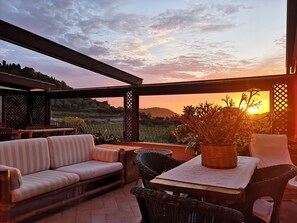A patio with a sofa, wicker chairs, and a table set for dining, overlooking a scenic landscape during sunset. - La Tinaia Country House (Magomadas)
