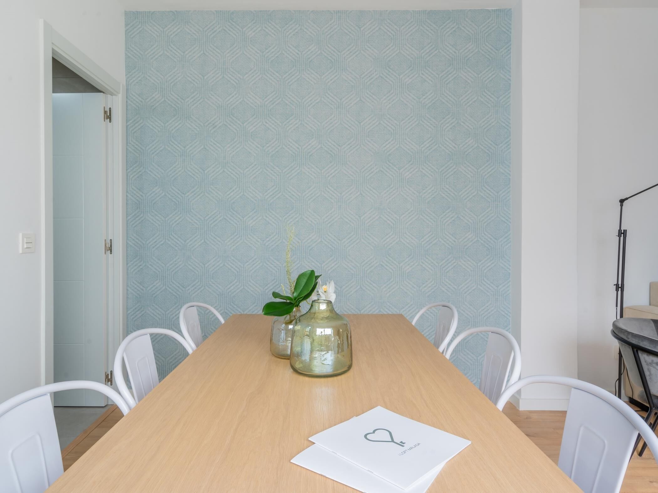 A dining area with a wooden table, white chairs, a patterned wall, and a small plant on the table.