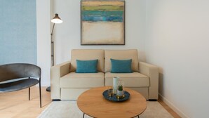 Un salon avec un canapé beige, deux coussins bleus, une table basse ronde en bois agrémentée d’objets décoratifs, un fauteuil noir et un lampadaire.