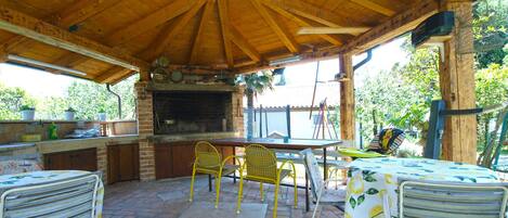 Una cocina al aire libre rústica con suelo de ladrillo, techo de madera y un horno de ladrillo. Hay dos sillas de metal y una mesa con mantel.