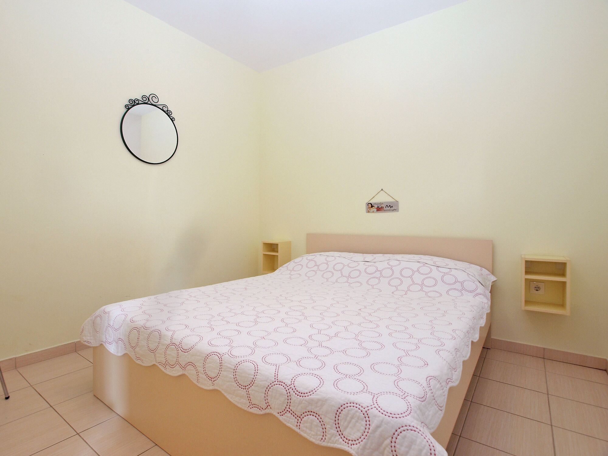 Een slaapkamer met een enkel bed, een nachtkastje, een wandspiegel en een klein plankje met een bordje.