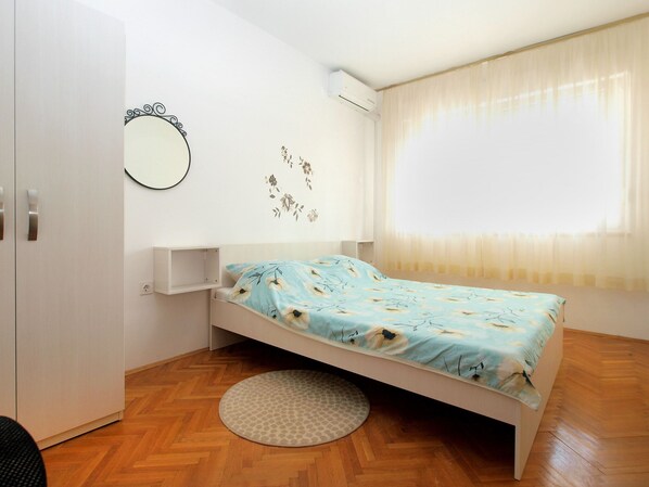 Un dormitorio con cama, un espejo redondo, una pequeña estantería y una ventana con cortinas.