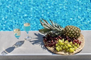 Cenário à beira da piscina com uma mesa exibindo uma abacaxi, uvas e dois copos de uma bebida azul clara.