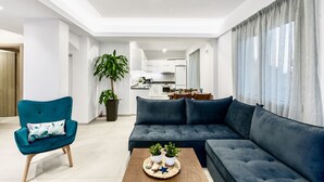 A modern living room with a dark blue sofa, a teal armchair, and a coffee table with decorative items. A large potted plant is in the corner, and the room has a neutral color palette. - Villa Izabela (Hersonissos)