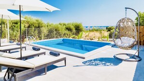 A swimming pool with lounge chairs and a hanging chair, surrounded by a fence and greenery. - Villa Izabela (Hersonissos)