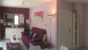 Een woonkamer met een paarse bank, een eettafel met stoelen, een klein wit tafeltje met een roze vaas en een vloerlamp.
