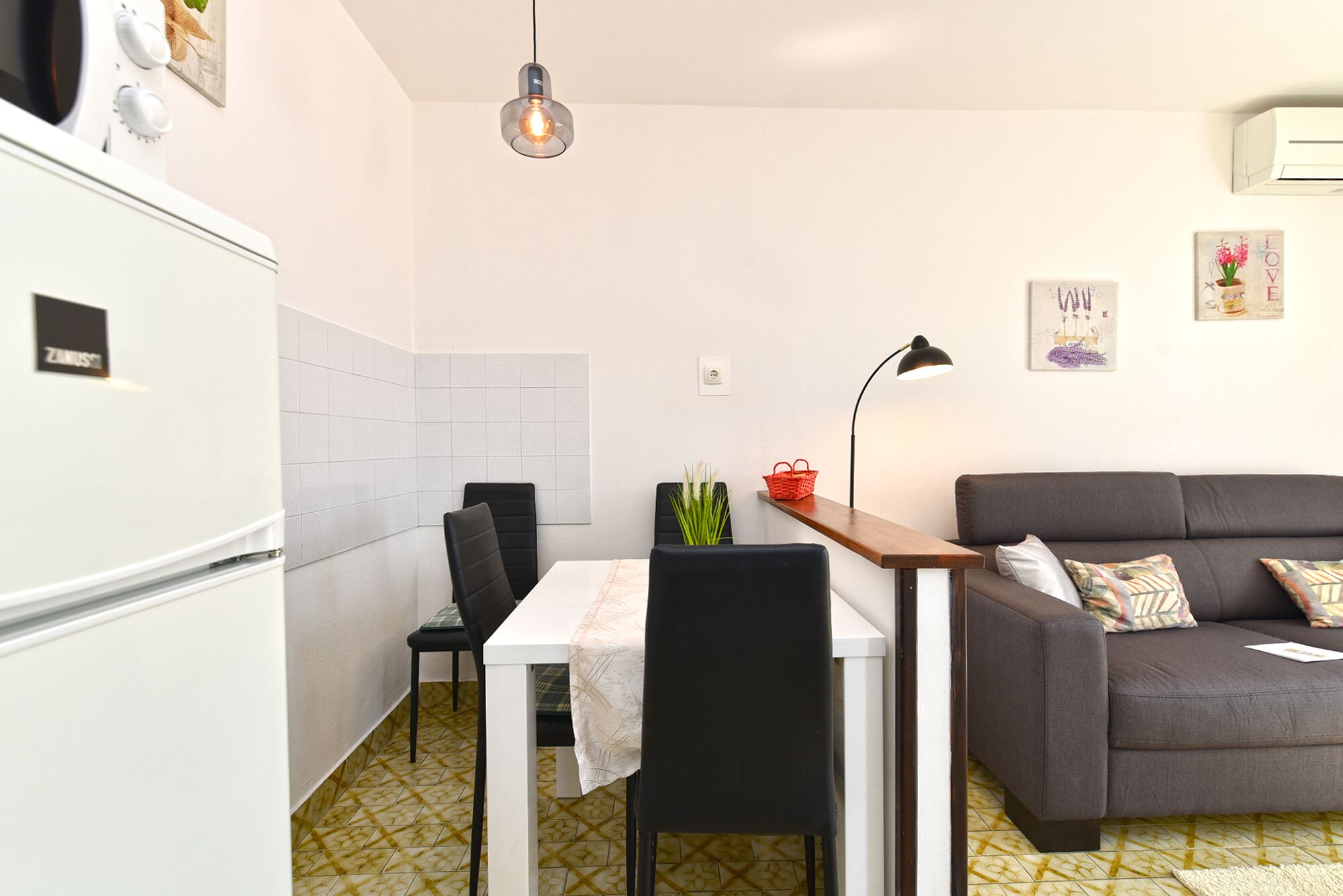 A compact kitchen with a refrigerator, a dining table with chairs, a sofa, and a small side table.