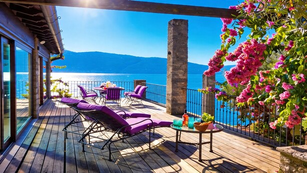 Eine Holzterrasse mit violetten Liegestühlen und einem Tisch, mit Blick auf ein Gewässer und Berge.