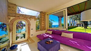 Ein Zimmer mit Steinwand, großen Fenstern mit Meerblick, einem violetten Sofa und einem kleinen Tisch mit Büchern.