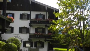 Un edificio de dos pisos con fachada blanca y contraventanas negras, rodeado por un césped bien cuidado y un columpio de madera.