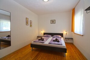 Ett sovrum med trägolv, en säng med lila sängkläder, två sänglampor och ett fönster med gardiner.