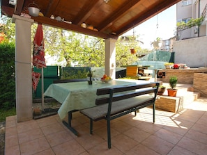 Un espace extérieur abrité, aménagé pour prendre les repas, avec une table et des bancs, entouré de plantes en pot et d’une clôture verte.