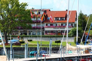 Un hôtel situé au bord de l’eau, avec un toit recouvert de tuiles rouges, des balcons ornés de fleurs et un port de plaisance rempli de bateaux.