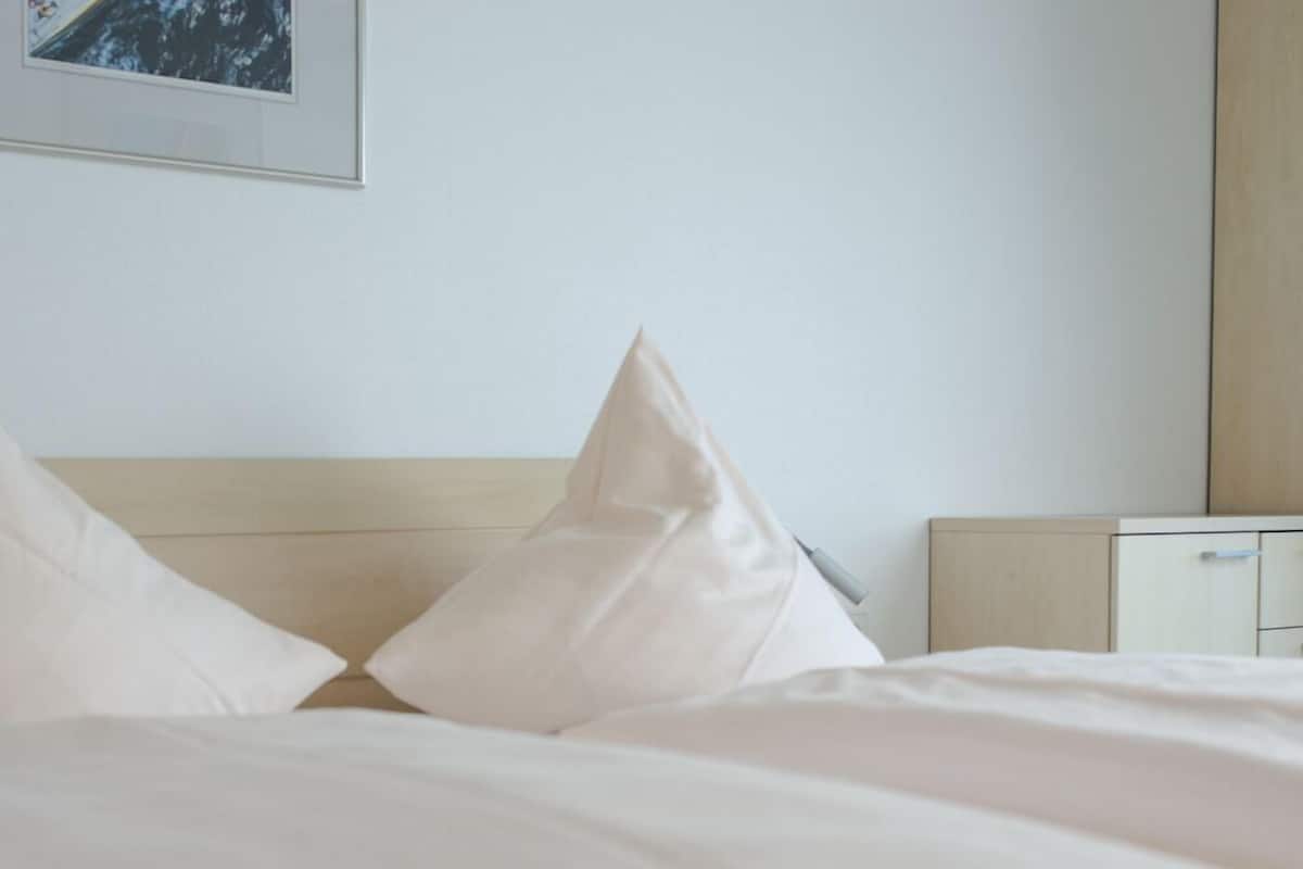 A neatly made bed with white linens and pillows, a wooden headboard, and a small bedside table with a lamp.