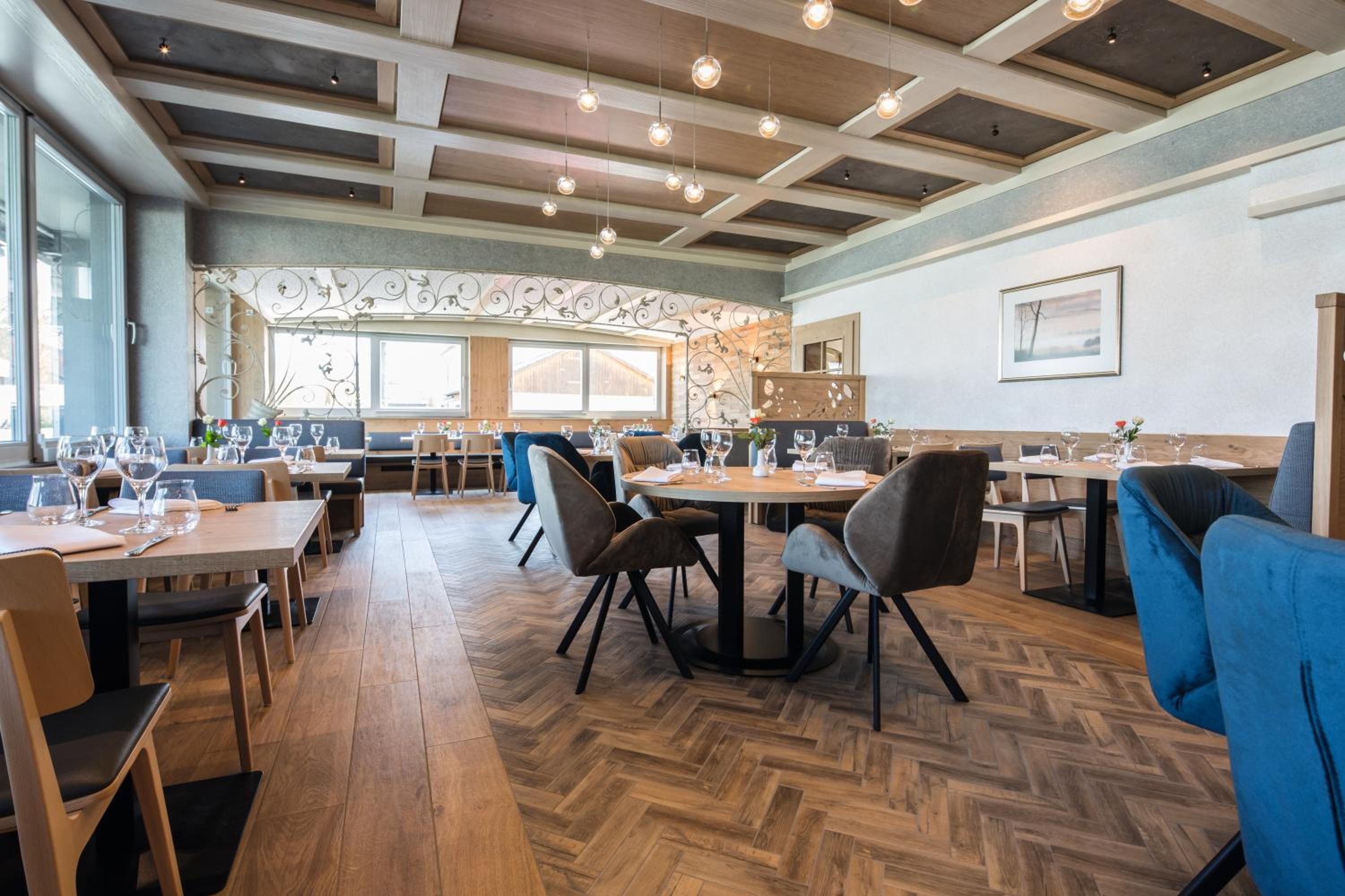 Interior de un restaurante con piso de madera, mesas, sillas e iluminación colgante.