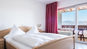 A hotel room with a wooden bed, white bedding, a nightstand, a red curtain, and a view of the city through a large window. - Landhotel Bodensee (Konstanz)