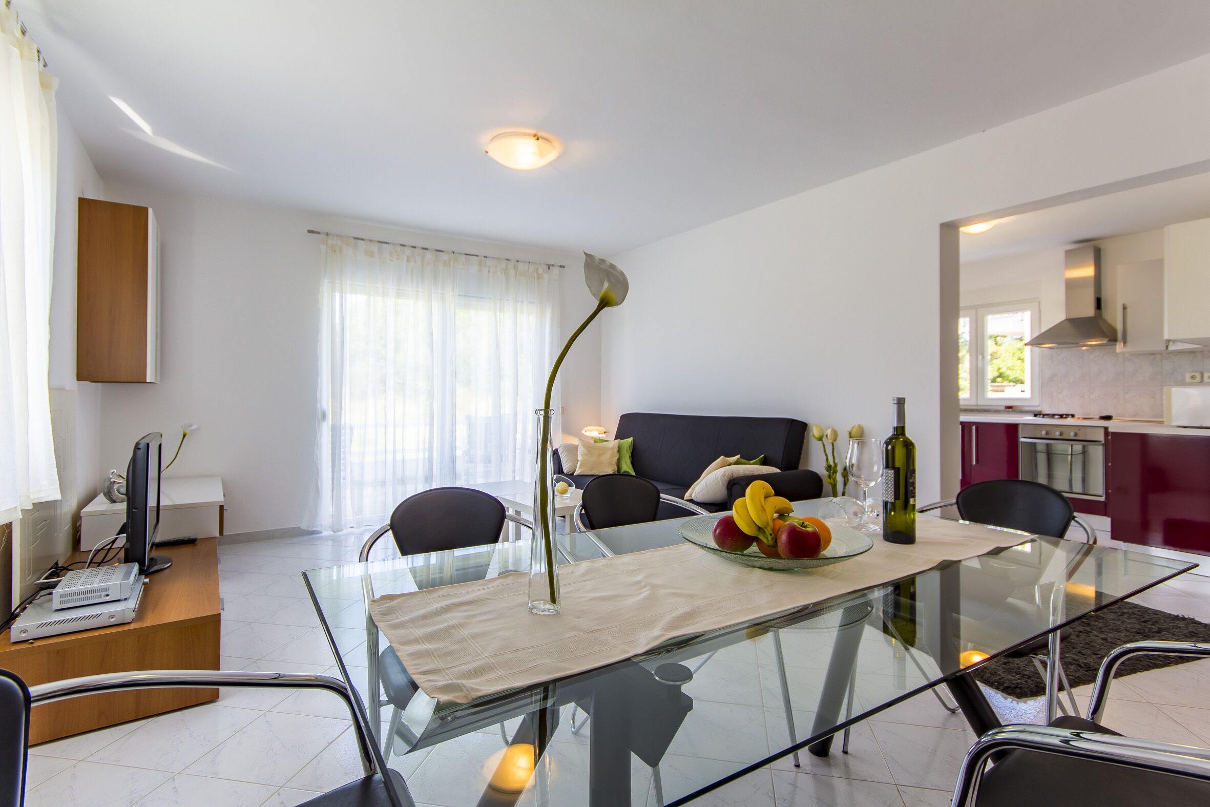 A modern living room with a glass dining table, a sofa, and a kitchen area in the background.