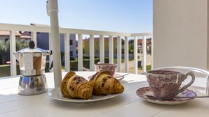 A table with a coffee maker, croissants, and a cup of coffee on a balcony with a view of buildings.
