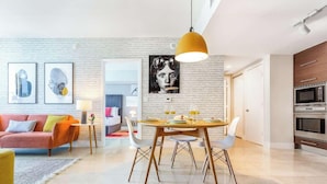 A modern living room with a sofa, dining table, and chairs, a kitchen with appliances, and a large wall art piece. - Brickell Urban Oasis | Modern Apartment (Miami)