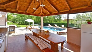 A covered outdoor area with a wooden ceiling, a pool, lounge chairs, and a dining table with cushions.