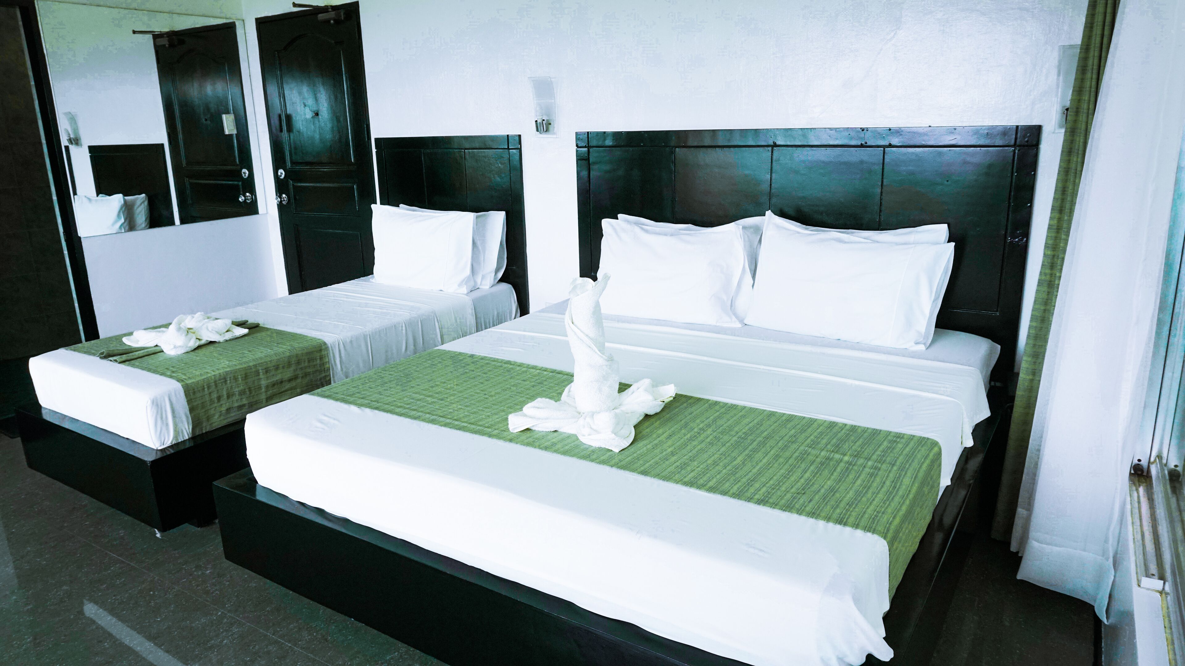 A hotel room with two beds, each with white linens and green bedspreads. There are two nightstands with lamps, a large mirror, and a door with a handle.