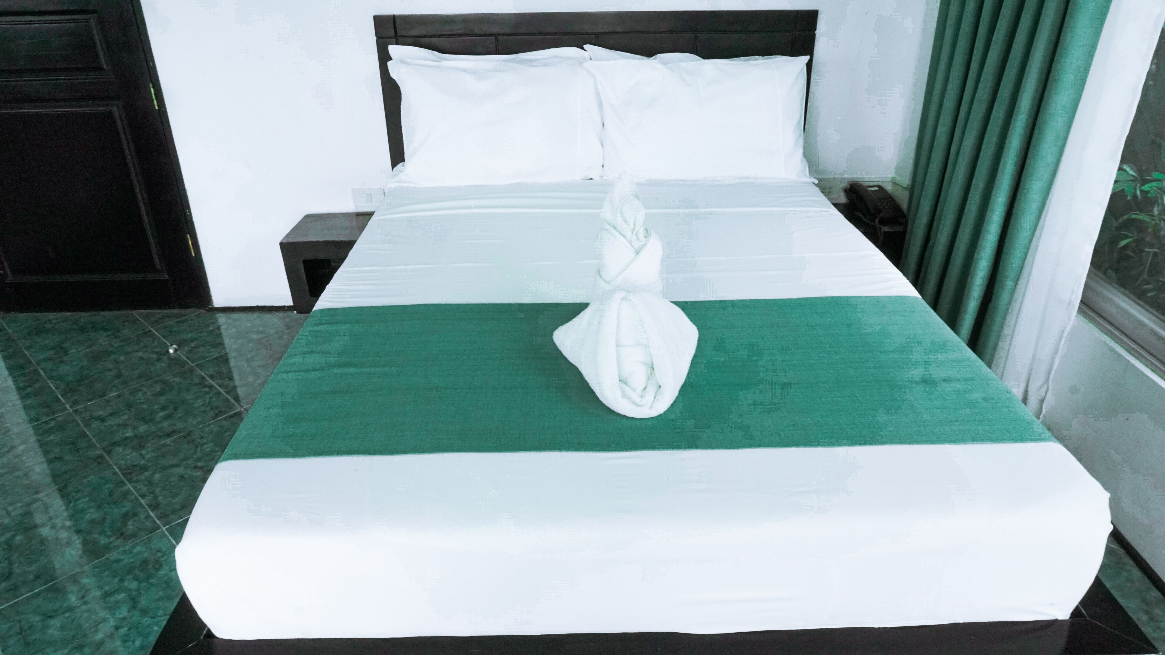 Un letto rifatto con lenzuola bianche e un tocco di verde, un comodino con un telefono e una finestra con tende.