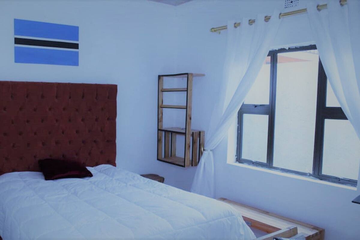 A bedroom with a single bed, a wooden nightstand, a small table, a window with curtains, and a flag on the wall.