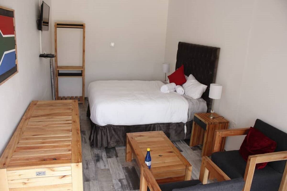A hotel room with a bed, a wooden desk, a chair, a small table, and a TV mounted on the wall.