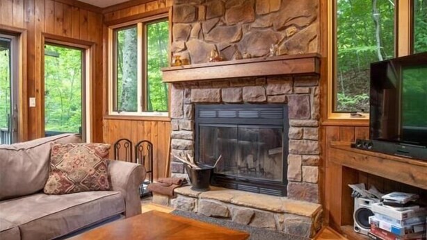 A cozy living room with a stone fireplace, wooden furniture, and large windows offering a view of the forest. - NF17- Cool, Creekside Home! Hot Tub! Pets ok! (Davis)