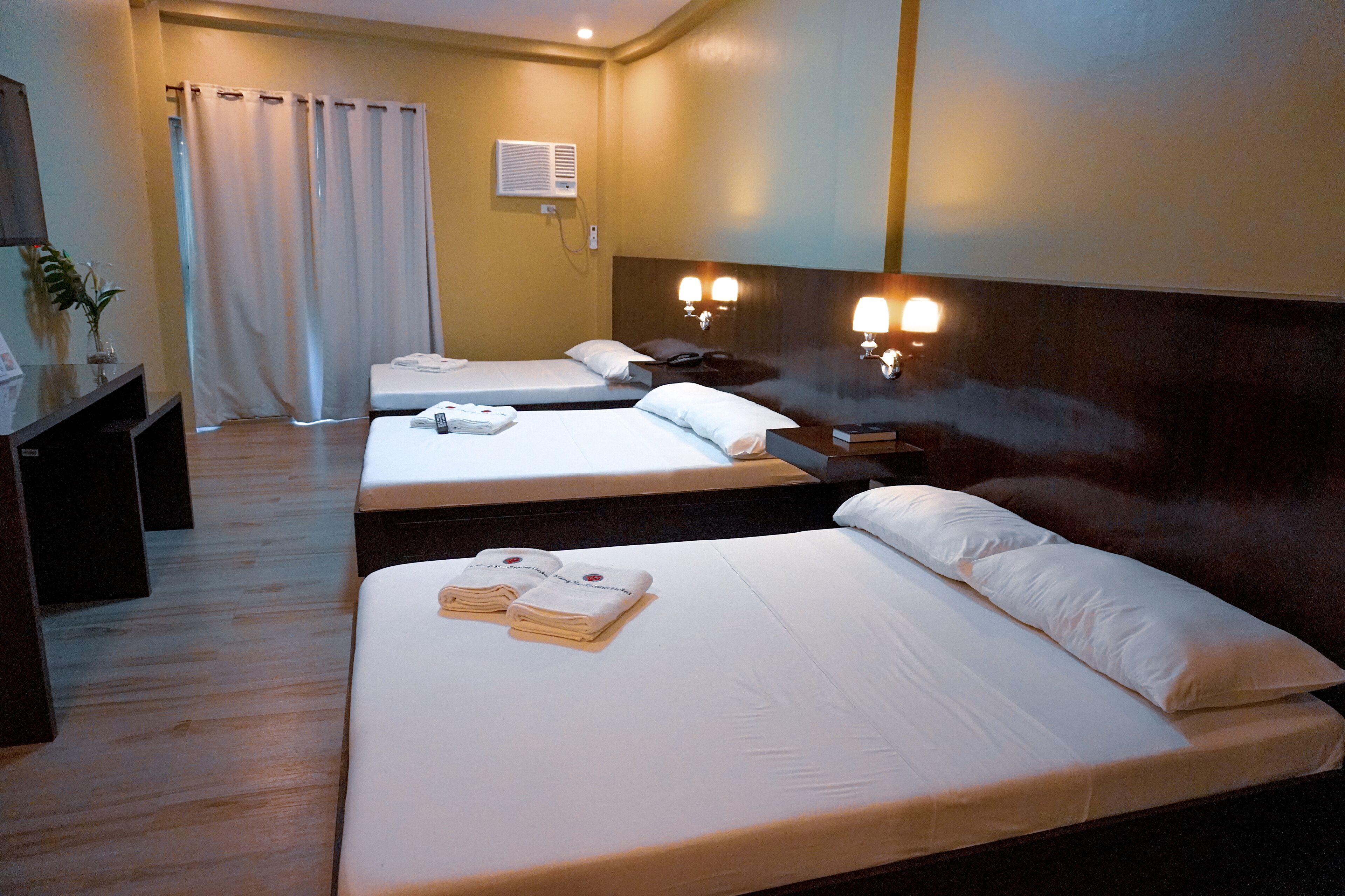 A hotel room with two beds, a desk, a chair, a plant, and a bathroom with a shower curtain.