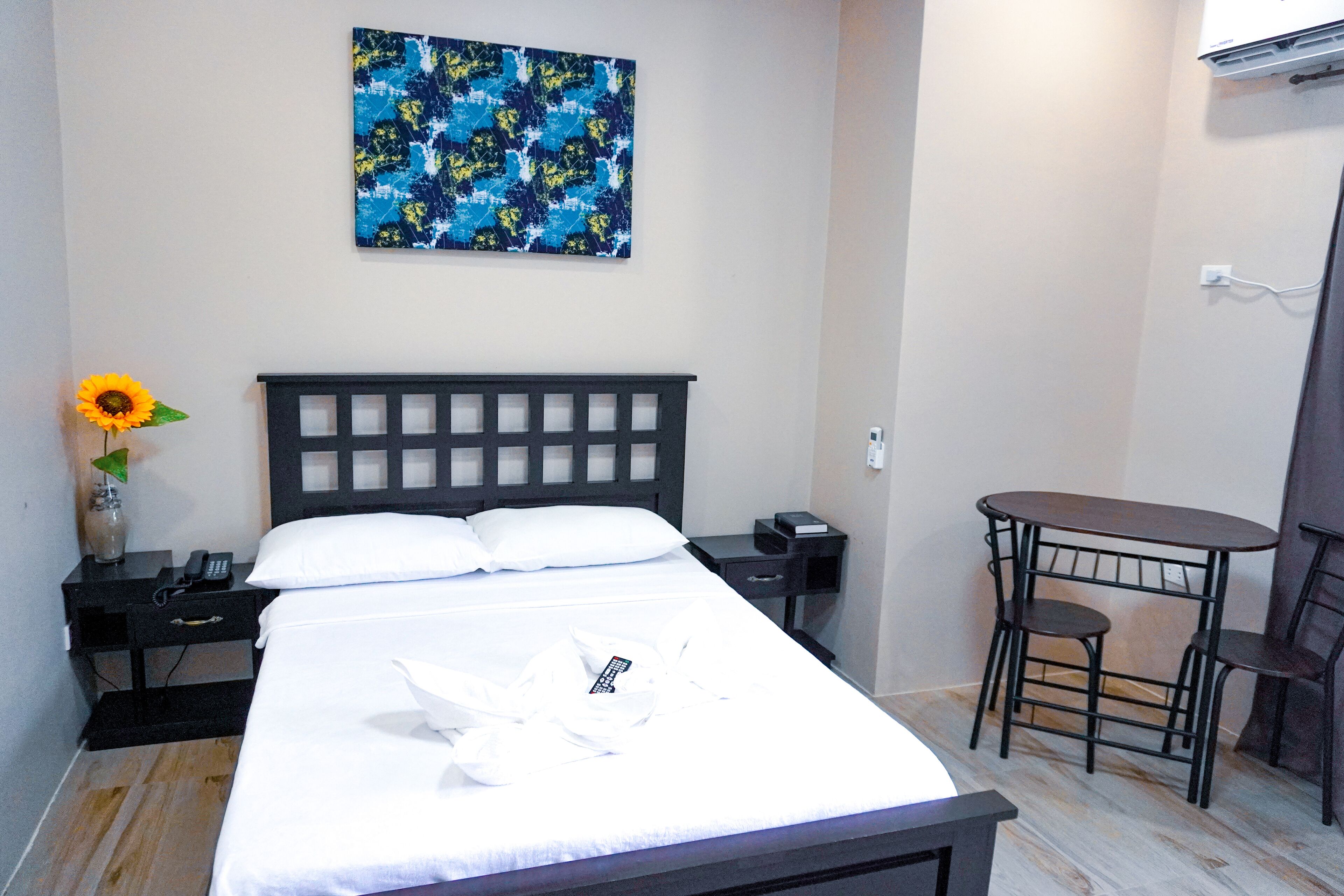 Un lit bien fait, avec du linge de lit blanc, une table de chevet avec un téléphone et un vase contenant un tournesol, une petite table avec des chaises et un climatiseur fixé au mur.