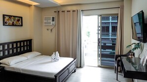 A hotel room with a bed, a desk, a TV, a window with curtains, and a wall-mounted air conditioner.