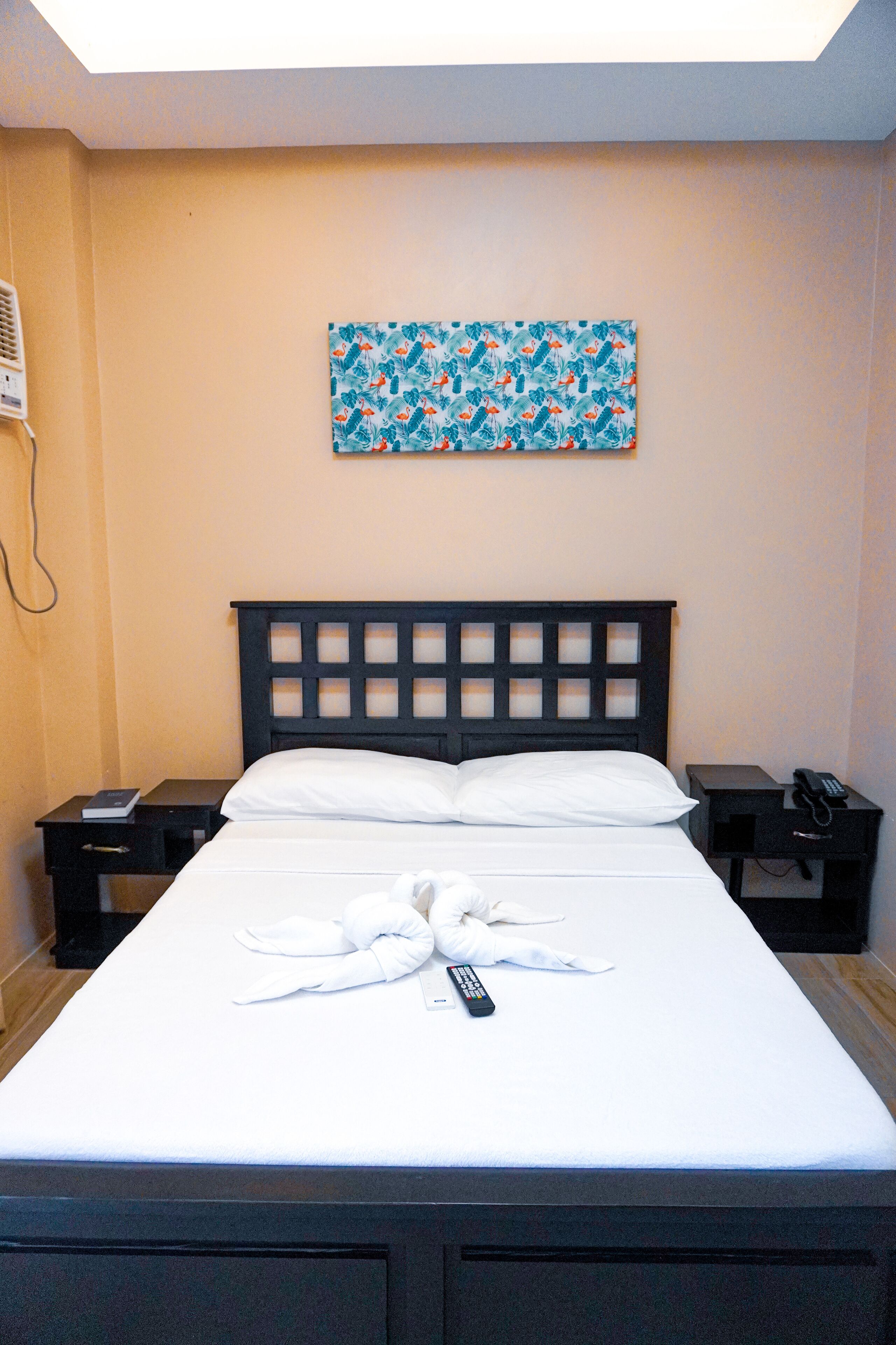 A neatly made bed with a remote control and a towel on it, a bedside table with a phone, and a painting on the wall.