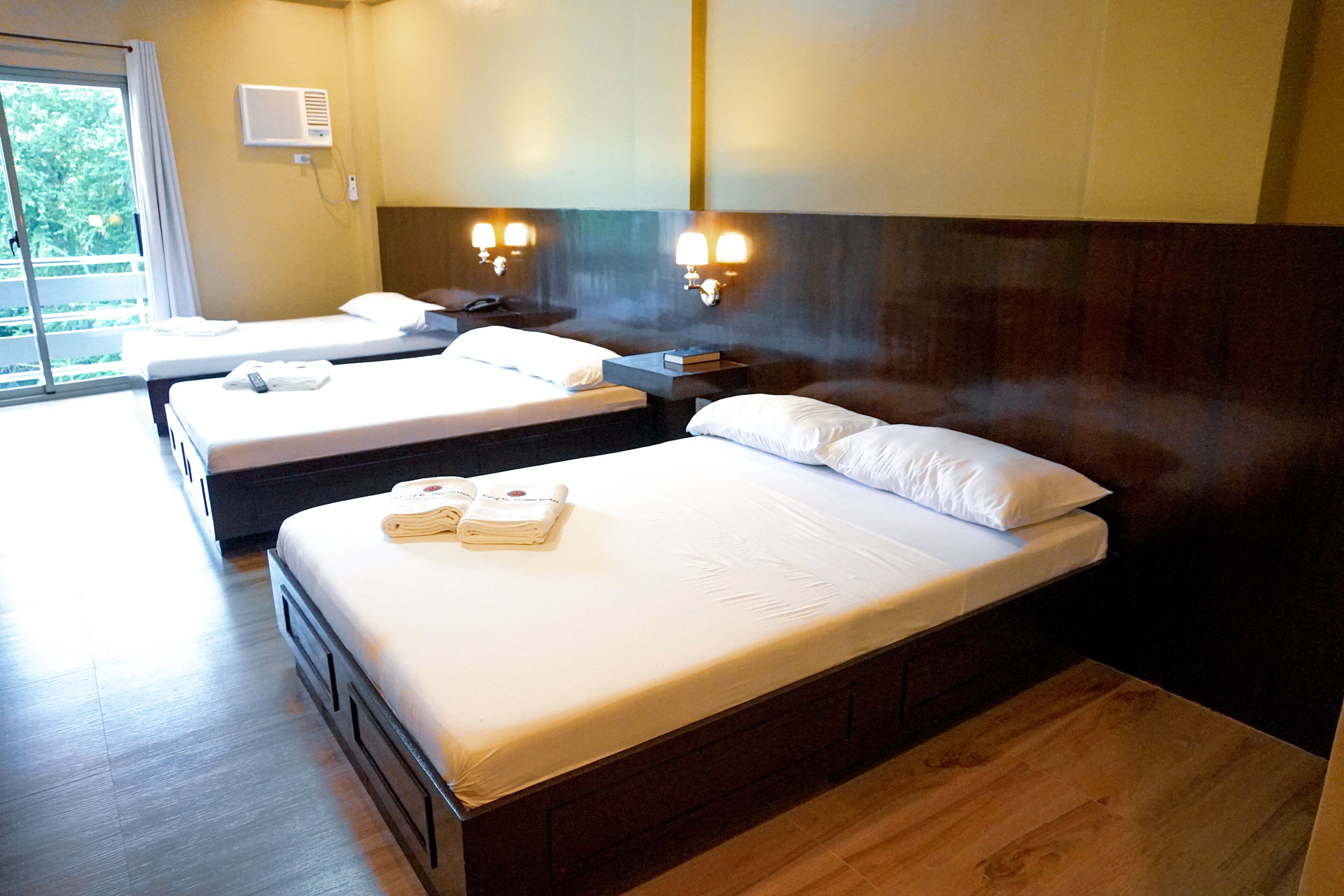 A hotel room with two single beds, a wooden headboard, a bedside table with a lamp, and a window with curtains.