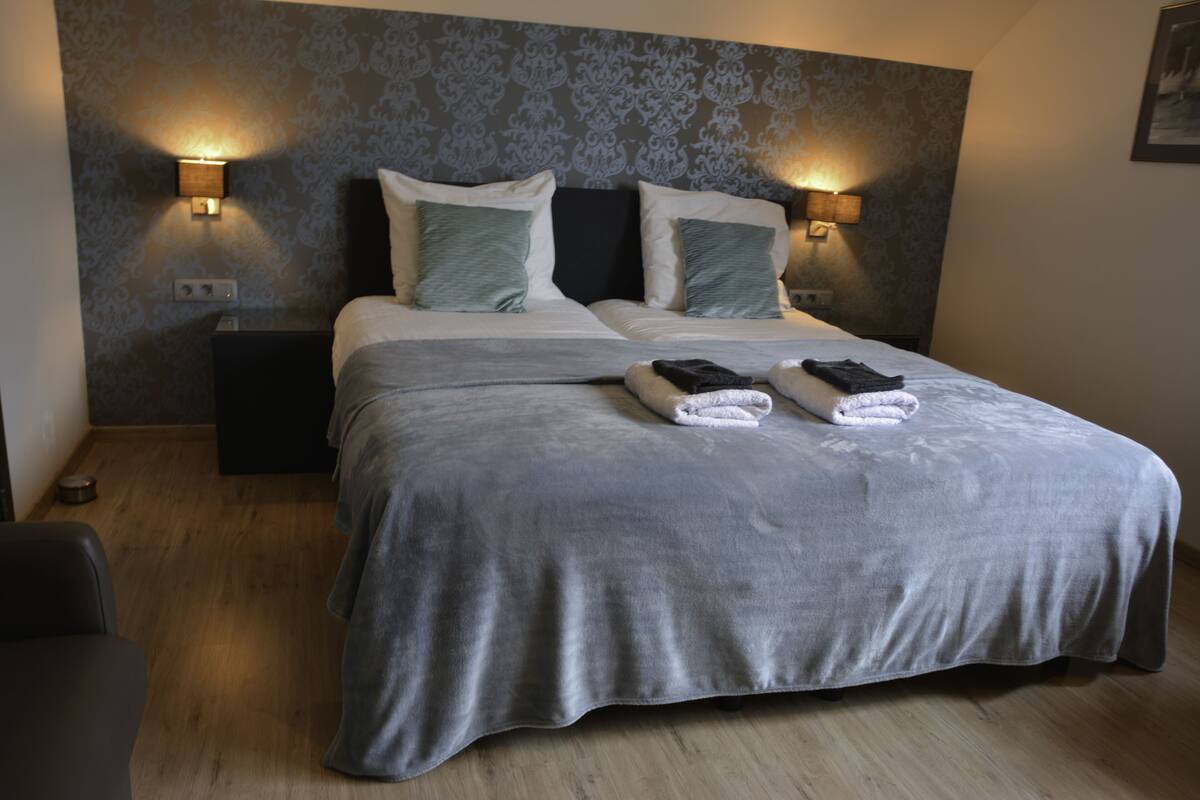 A neatly made bed with a grey bedspread and two pillows, a bedside table with a lamp, and a wall with a patterned wallpaper.