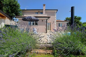 A two-story house with a stone facade, a tiled roof, and a swimming pool in the background.