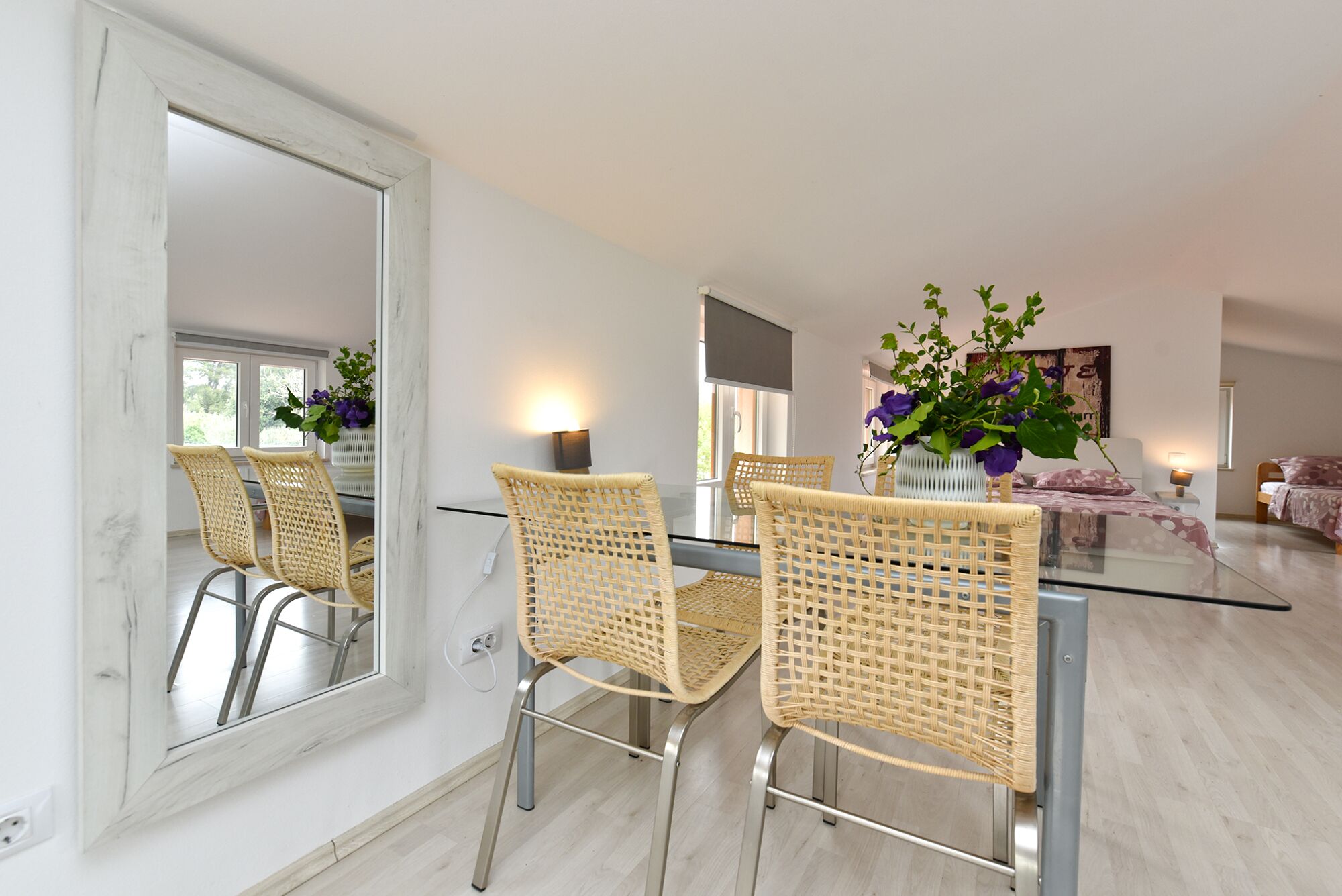 A dining area with wicker chairs, a glass table, and a large mirror.