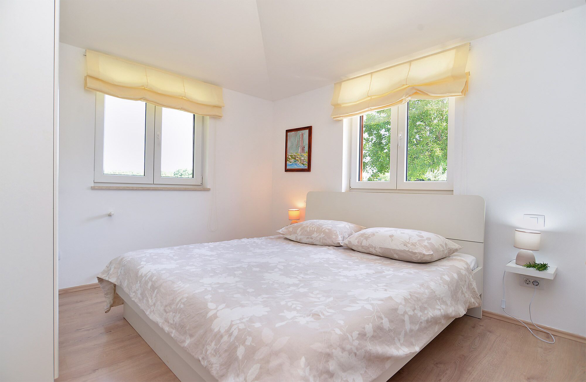 Ein Schlafzimmer mit einem Bett, zwei Fenstern mit Vorhängen, einer Nachttischlampe und einem kleinen Regal mit einer Pflanze.