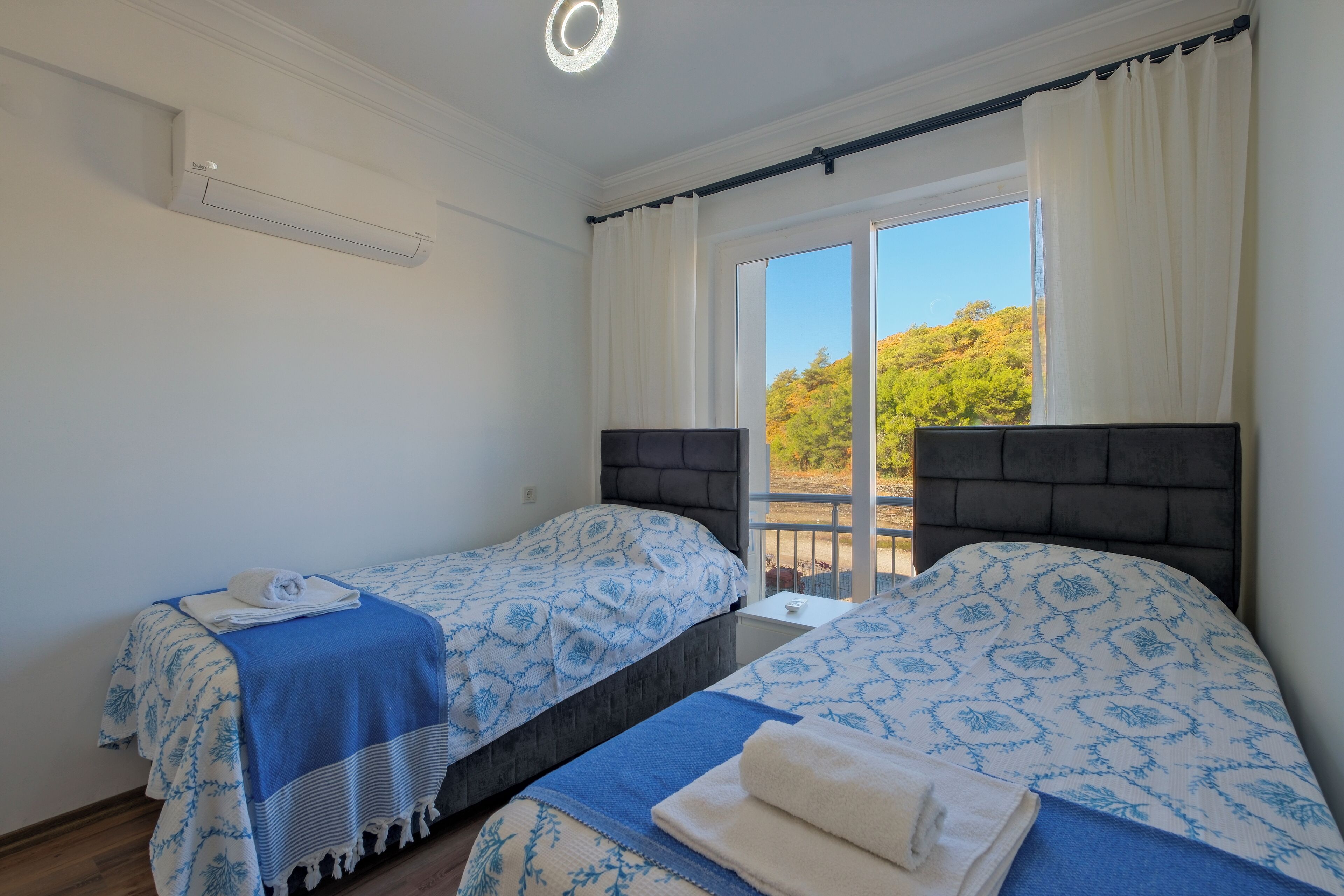 Une chambre avec deux lits, une fenêtre avec des rideaux, un luminaire au plafond et une vue sur la verdure.