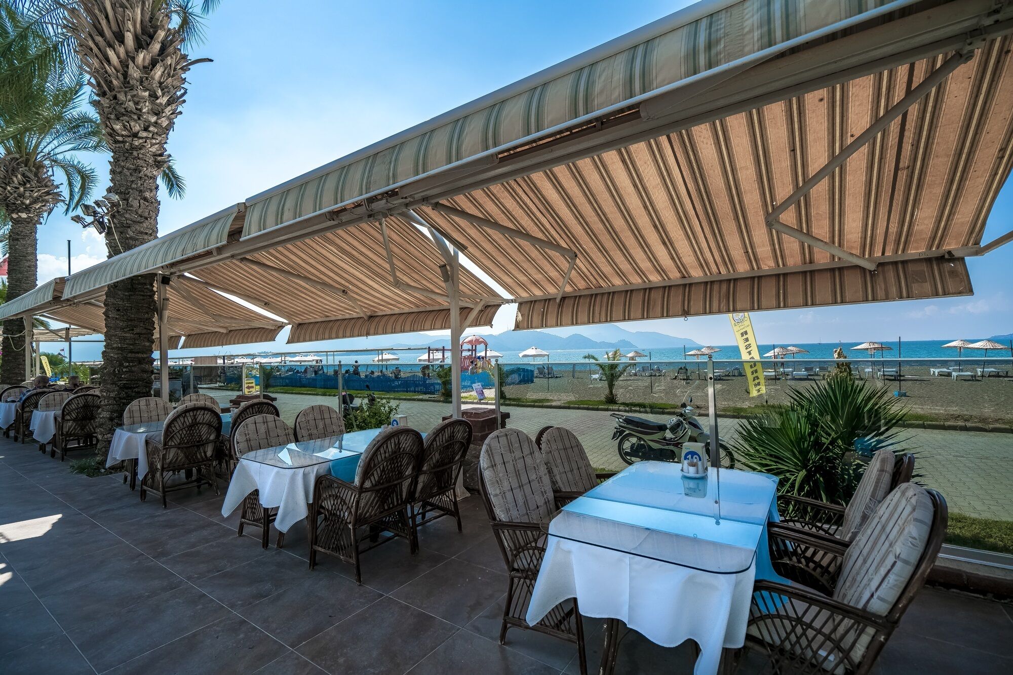 Un restaurant en plein air, avec des tables et des chaises disposées sous une tonnelle ombragée, offrant une vue sur une plage parsemée de parasols et de bateaux.