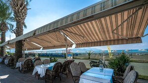 Un restaurant en plein air, avec des tables et des chaises disposées sous une tonnelle ombragée, offrant une vue sur une plage parsemée de parasols et de bateaux.